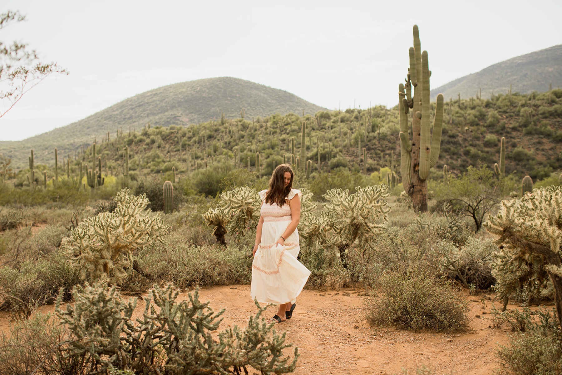 Heather Flynn in a white dress walking through the desert among saguaro cacti and distant mountains